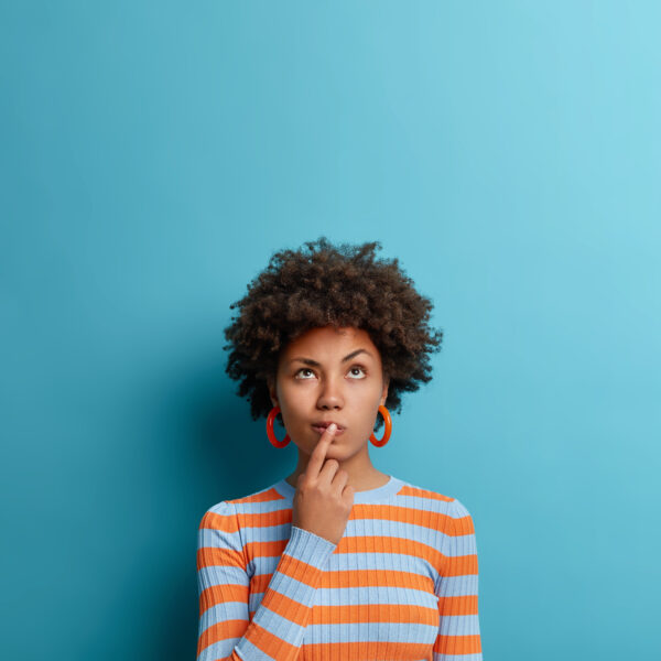 Vertical shot of thoughtful dark skinned woman has idea in mind, keeps finger on lips, looks pensively above, tries to decide how to act, poses against blue background, blank copy space above